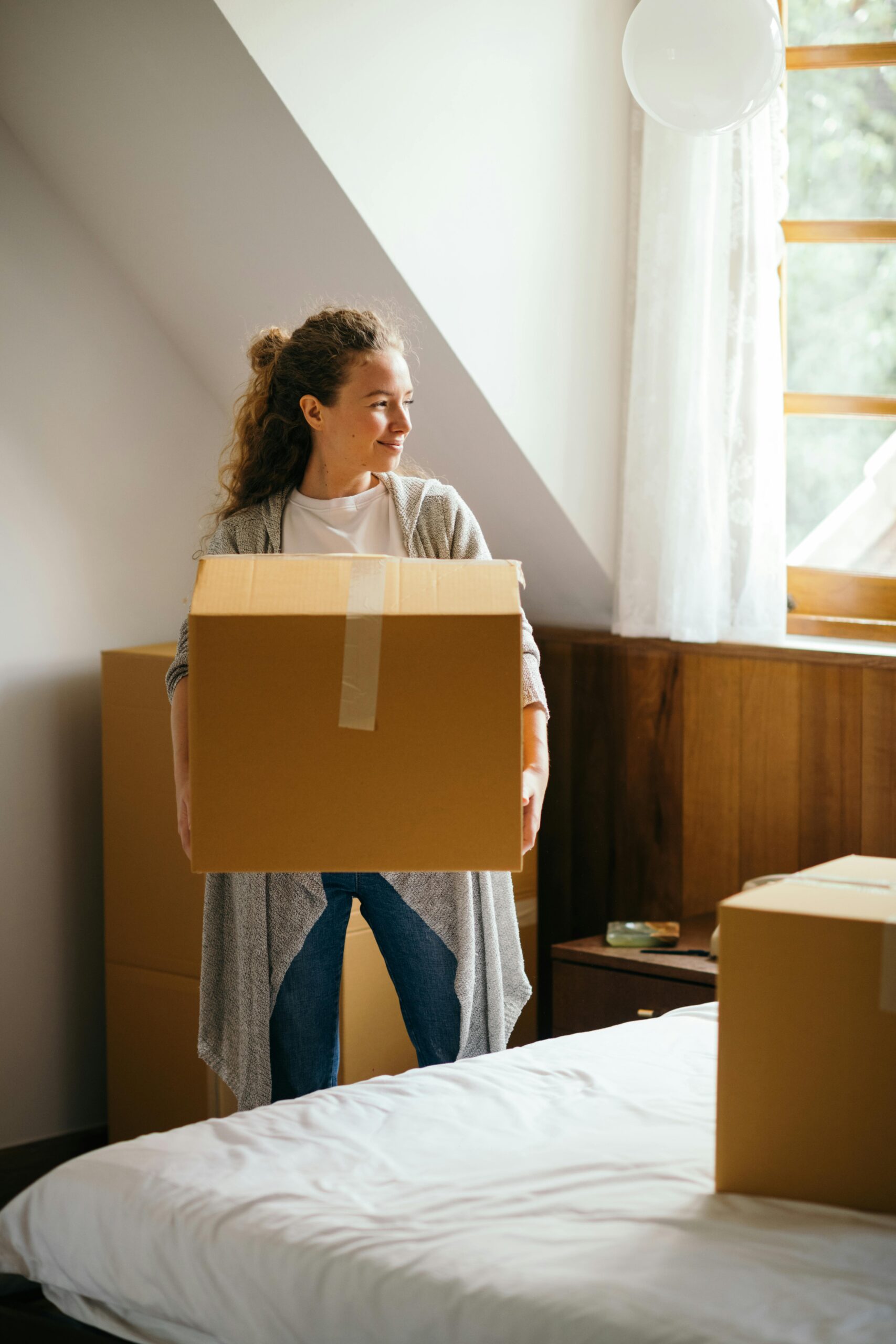 Young smiling lady in casual outfit with big cardboard box in hands standing and looking away near bed in modern bedroom of newly purchased house