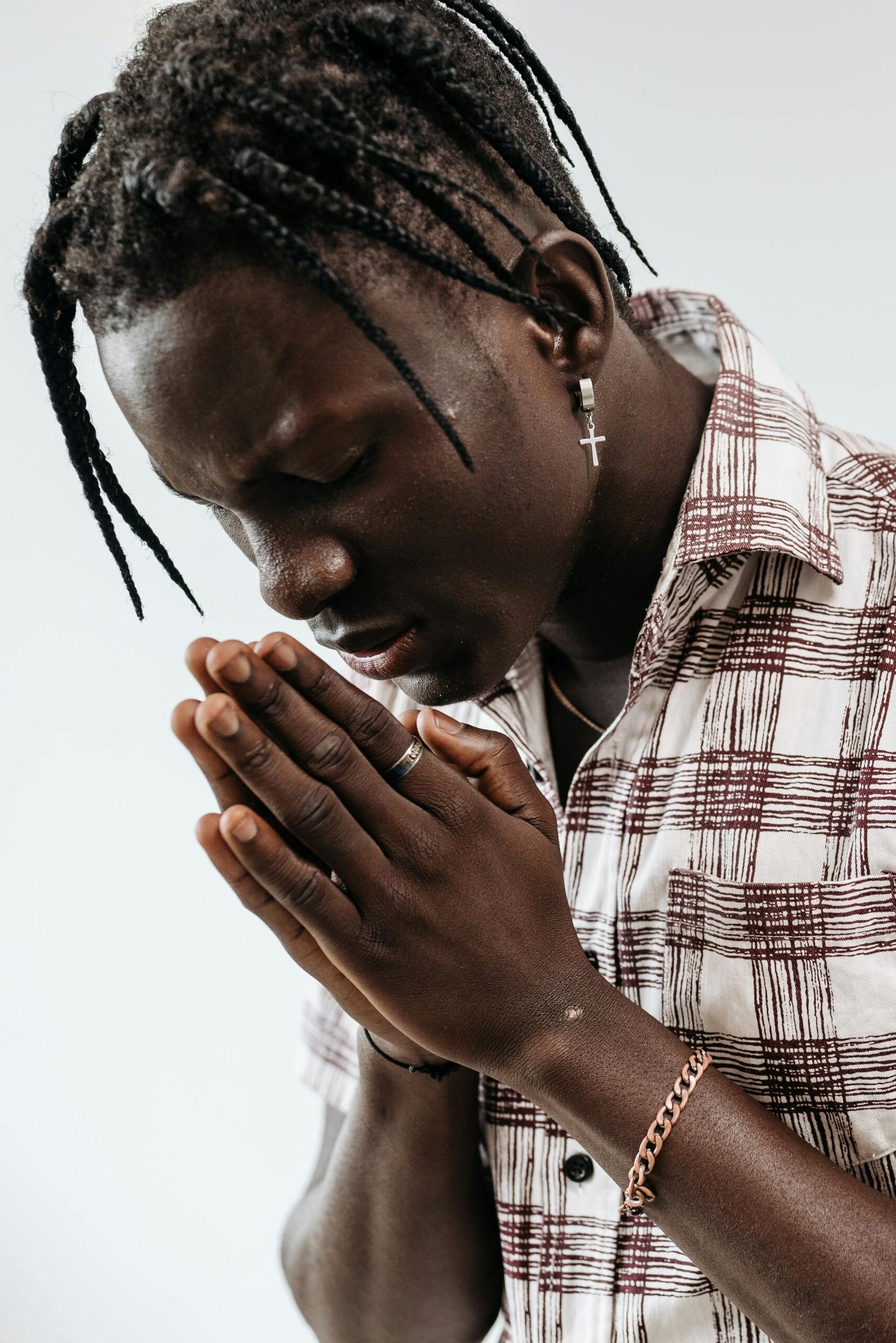 Side view of a young black man with braided hair in prayer, eyes closed, wearing a plaid shirt and cross earring.