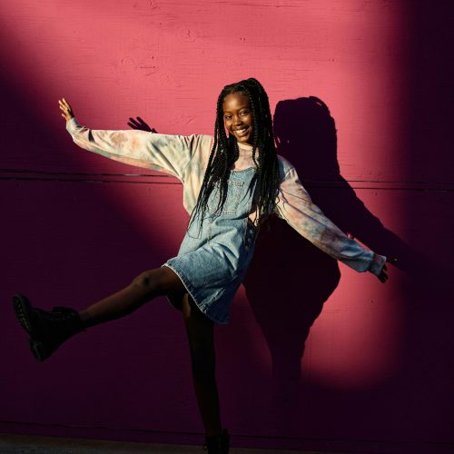 pexels-photo-23710190-23710190 Smiling teenage girl in denim dress posing playfully against a vibrant pink wall.